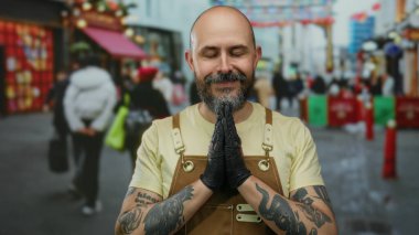 Dövmeli, kolları ve sakalı olan kel bir adam, şehir sokaklarında elleri sımsıkı bağlı, kahverengi önlük ve siyah eldivenlerle meditasyon yapıyor..