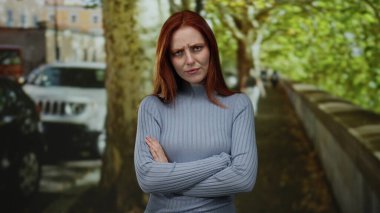 Kızıl saçlı bir kadın ağaçlarla kaplı bir şehir sokağında dikiliyor. Etrafı yeşilliklerle ve park edilmiş arabalarla çevrili, üzerinde şık bir süveter olan güzel ve düşünceli bir kadın..