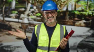 Güvenlik yeleği giymiş orta yaşlı bir adam açık şehir parkında elinde pano tutarken gülümsüyor..