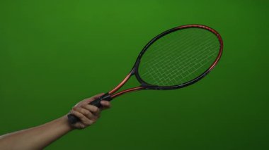 Erkek olan tenis raketini sıkı bir yeşil duvar arka planında tutuyor, spor malzemelerini gösteriyor ve koluna ve raketine odaklanıyor..