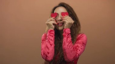 Gözlerinin üzerinde kırmızı kalp kesikleri olan ve gülümseyen bir kadın, şeftali arkaplanlı eller stüdyoda yüze yakın, neşeli romantizm.