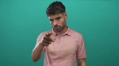 Pembe polo tişört giyen genç İspanyol adam yeşil bir stüdyoda parmağını doğrudan kameraya doğrultuyor..