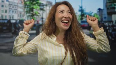 Uzun kahverengi saçlı, çizgili tişört giyen, yoğun bir şehir sokağında gün boyunca yumruklarıyla gülümseyen genç bir kadın..