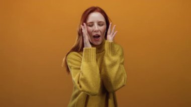 Turuncu arka plan önünde gözleri kapalı bağıran, rahat bir süveter giyen, heyecanlı ya da sürpriz yapan, neşe ve duygu içeren kızıl saçlı, kadın varlığını vurgulayan bir kadın..