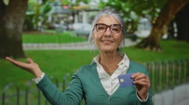 Parktaki son sınıf öğrencisi kadın elinde kredi kartıyla güneşli bir günde açık havada neşe saçıyor, gözlük takıyor ve yeşil hırka giyiyor..
