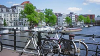 Bisikletler, güneşli bir günde sakin suyun yanında geleneksel Hollandalı binaların bulanık arka planlarıyla Amsterdam 'da bir kanal korkuluğuna dayanır..