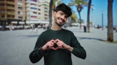 Genç adam güneşli bir şehir sokağında yeşil bir kazak giymiş, kameraya gülümseyen, sıcak ve misafirperver bir atmosfer yaratan bir kalp hareketi yapıyor..