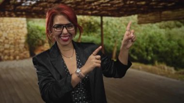 Kadın verandada işaret parmağı yukarı doğru, gözlüklerle ve katmanlı kolyelerle gülümsüyor; keyif güveni.