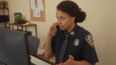 Üniformalı kadın polis masasında telefonla konuşuyor, bilgisayar ve evrak işleriyle meşgul..