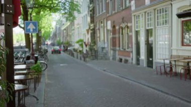 Ağaçlarla kaplı, bisikletli dar bir yolun yanında sakin bir açık hava kafe, klasik mimari ve şehir cazibesini yumuşak odak noktasında sergileyen Avrupa sokak manzarası.