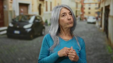 Arka planda kaldırım taşları ve bulanık binaları olan bir sokakta dikilirken parmaklarına güvenen yaşlı bir kadın..