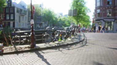 Motosikletli, güneşli ağaçlı ve kentsel yaşamı yansıtan tarihi binalı bir kanalda yürüyen insanların bulanık görüntüsü.