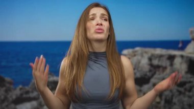 Kadın omuzlarını silker ve stüdyoda parlak güneş ışığı altında kayalık deniz manzarasına karşı ellerini kaldırır..