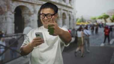 Akıllı telefon tutan genç Çinli adam güneş ışığı altında Roma Kolezyumu 'nun girişinde olumlu bir hareket sergiliyor..
