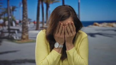 Güneşli deniz kıyısında palmiye ağaçlarıyla gezerken, yüzünü endişeye boğan bir kadın..