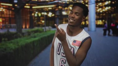 ABD basketbol forması giyen bir adam sokakta çitlerle ve şehir ışıklarıyla kaplı bir objeyi işaret ediyor..