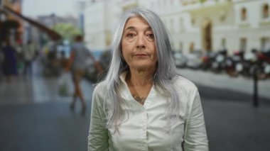 Beyaz tişörtlü, hareketli bir caddede duran gri saçlı yaşlı kadın yoğun şehir geçmişine uyum sağlıyor..