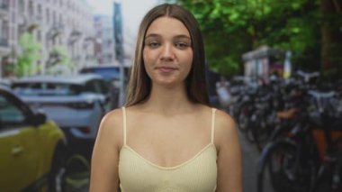 Kadın, park halindeki bisiklet ve arabalarla çevrili işlek şehir caddesinde kaldırım kenarındaki ağaçların ve mağazaların önünde el kaldırıyor..