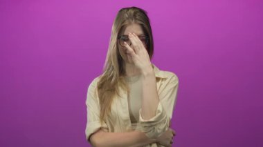 Sarışın kadın, alnına dokunarak gözlük takıyor ve pembe stüdyoda kollarını çapraz yapıyor. Utanç verici..