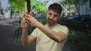 Latin kökenli bej tişört giyen genç bir adam, binaları ve yeşillikleri olan güneşli bir caddede bisiklet önünde parmaklarını birbirine geçiriyor..