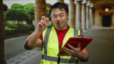 Güvenlik yeleği giymiş bir adam üniversite avlusunda panoyla poz veriyor, akademik ortamı tarihsel mimari ile çevrili günlük teftiş temasıyla harmanlıyor..