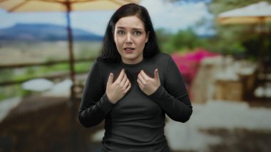 Esmer bir kadın, güneşli bir restoranın terasında, arka planda dağlar olan manzaralı bir caddeye bakarak anlamlı bir şekilde el kol hareketi yapıyor, duygularını iletiyor..