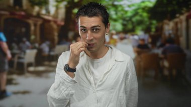 Beyaz gömlekli genç adam sessiz güneşli sokak restoranının terasında sessizlik için parmaklarını dudaklarına bastırıyor..