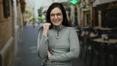 Bir şehir caddesinde gözleri kapalı gülümseyen bir kadın, esmer saçı ve gözlüğü açık kafelerin arka planına vurgulanmış ve bulanık yayalar.