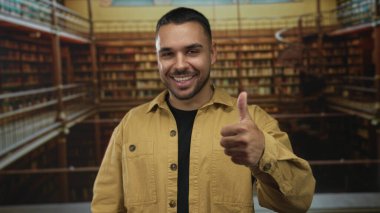 Genç İspanyol gülümseyen adam, ahşap raflarla kaplı eski kütüphane binasında baş parmak hareketi gösteriyor. İyimserlik..