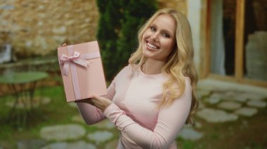 Açık hava restoranında elinde pembe bir hediye tutan genç bir kadın masaları ve taş yolları olan, neşeyle gülümseyen bir kadın..