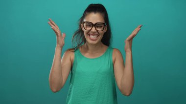 Latin kökenli genç bir kadın el kaldırıyor ve yüzünü buruşturuyor, gözlük takıyor ve yeşil kolsuz bluz giyiyor..