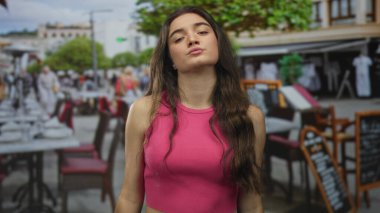 Çıplak omuzlu genç İspanyol kadın açık hava yemek masalarıyla çevrili güneşli restoranın terasında kulaklarının arkasına saçlarını örten pembe bir atlet giyiyor..