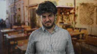 Açık hava restoranının terasında sakallı genç bir adam çizgili gömlekli canlı bir şehir ortamında hoşnutsuz görünüyor..