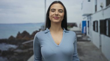 Arka planda okyanus ve gökyüzüyle deniz kenarında gülümseyen bir kadın. Sahildeki sahnenin güzelliğini vurgulayan neşeli bir ifade..