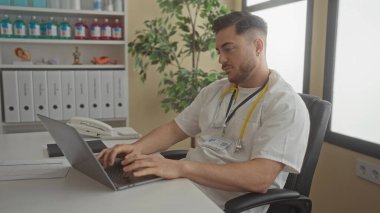 Sakallı ve steteskoplu genç bir doktor tıbbi malzeme ve ciltlerle dolu modern bir hastane odasında dizüstü bilgisayarla çalışıyor..