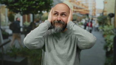 Sakallı kel adam şehir merkezinde kulaklarını kapatıyor. Gri süveter giyiyor. Şehir ortamında gürültü ya da konsantrasyon öneriyor..