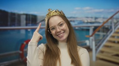 Beyaz gözlüklü genç bir kadın açık gökyüzünün altında güvertede taçtan tacı işaret ediyor; kraliyet güveni.