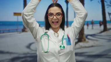 Steteskoplu kadın doktor, deniz kenarında palmiye ağaçlarıyla dolu bir yerde kalp jesti yapıyor. Üniformayla dışarıdaki sükuneti kucaklıyor..