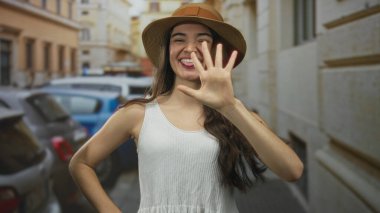 Hasır şapkalı ve beyaz kolsuz tişörtlü kadın dar bir caddede gülümserken beş parmağını kaldırıyor, dikkat çekiyor; oyuncu sayma.