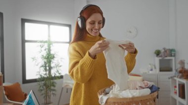 Kulaklık takan bir kadın, rahat bir odada çamaşırlarını sıralıyor sarı bir kazak giymiş bir sepet elbise ve bir bitki taşıyor..