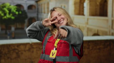 Sarı saçlı, kırmızı yelekli, gülümseyen ve dışarıdaki üniversite bahçesini gösteren genç bir kadın gönüllü..