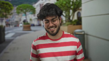 Sakallı genç adam canlı bir sokak ortamında gülümsüyor, gözlük takıyor ve kırmızı çizgili bir gömlek giyiyor, mutluluk ve rahatlama taşıyor..