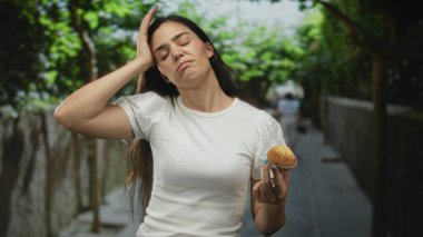 Yaz yeşilliğinin ortasında yapraklı ağaçlarla kaplı, güneşli bir sokakta kadın kek tutuyor ve alnına dokunuyor..