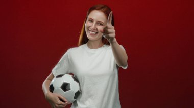 Kırmızı arka planda futbol topunu tutan kadın, işaret eden bir hareketle güvenle gülümsüyor..