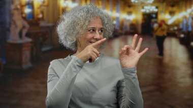 Gri saçlı bir kadın müzede el hareketi yapıyor, gülümsüyor ve sofistike bir iç mekan ortamında pozitifliği simgeliyor..