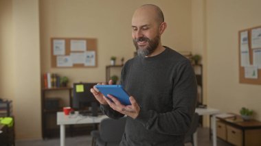 Ofiste sakallı bir adam tablet kullanıyor. Etrafında bitkiler ve bilgisayar ekipmanları var.