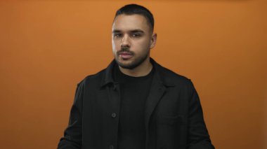 Adam açık avuç içleri ile ellerini uzatır ve portakal renkli stüdyo ortamında düşünceli bir bakış tutar; sakin yansıma ve odaklanma.