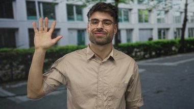 Bej gömlekli ve gözlüklü bir adam güneşli ağaçlı caddede selamlaşırken el kaldırıyor..