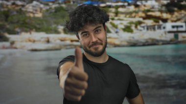 Genç sakallı bir adam, berrak mavi bir deniz ve kayalık bir kıyı şeridi ile manzaralı bir plaj zemininde olumlu ve neşeli bir hava yayıyor..