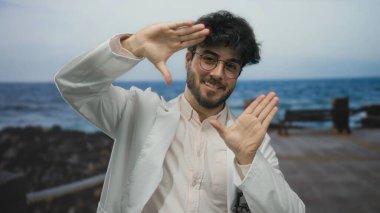 Sakallı ve gözlüklü genç adam güneşli bir günde arka planda deniz kenarında el kol hareketleri yapıyor.